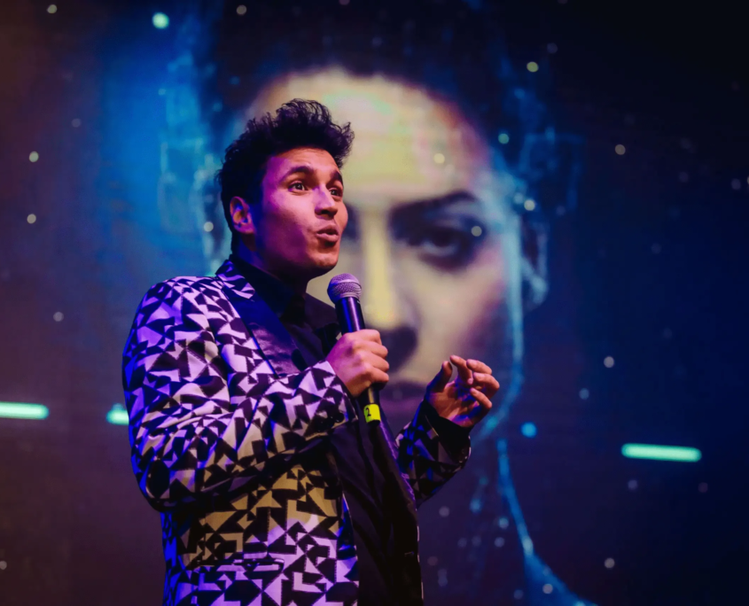 A performer on stage holding a microphone with face obscured by privacy block, wearing a geometric-patterned jacket under blue stage lighting.