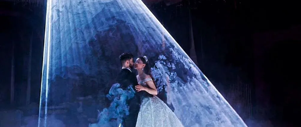 A couple in a spotlight on a stage with ethereal decorations, sharing a romantic moment.