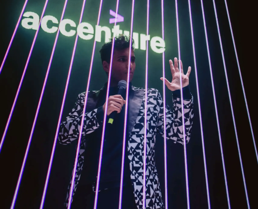 : Pessoa com sinal de paz em frente à iluminação e feixes de luz "Accenture"