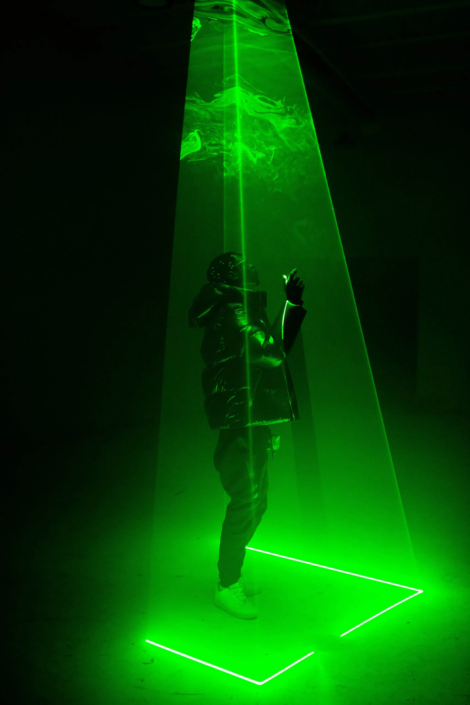 Uma figura em um traje espacial está em uma coluna triangular de luz verde.