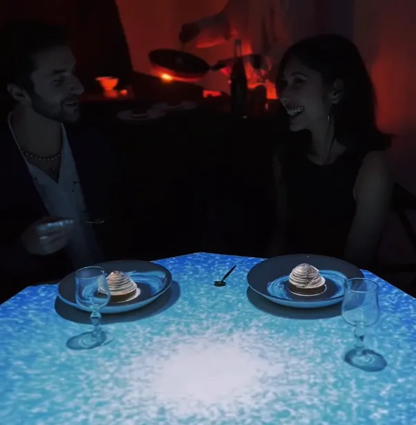 Um casal janta em um ambiente imersivo com a mesa iluminada por projeções digitais.