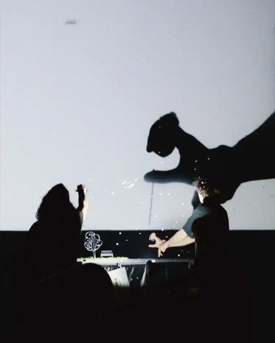 Artistas criando silhuetas de sombras em uma tela de projeção gigante durante um espetáculo digital narrativo.