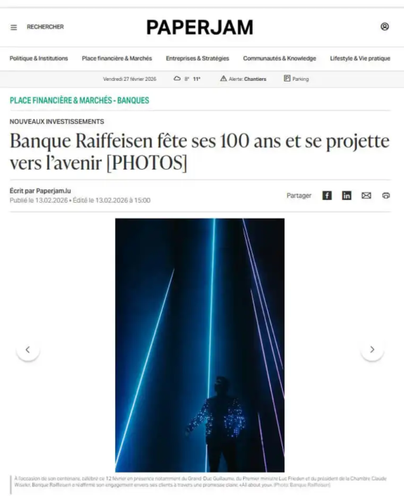 Skærmbillede af en nyhedsartikel fra Paperjam med titlen "Banque Raiffeisen fejrer 100 år og ser mod fremtiden", illustreret med et lasershow.